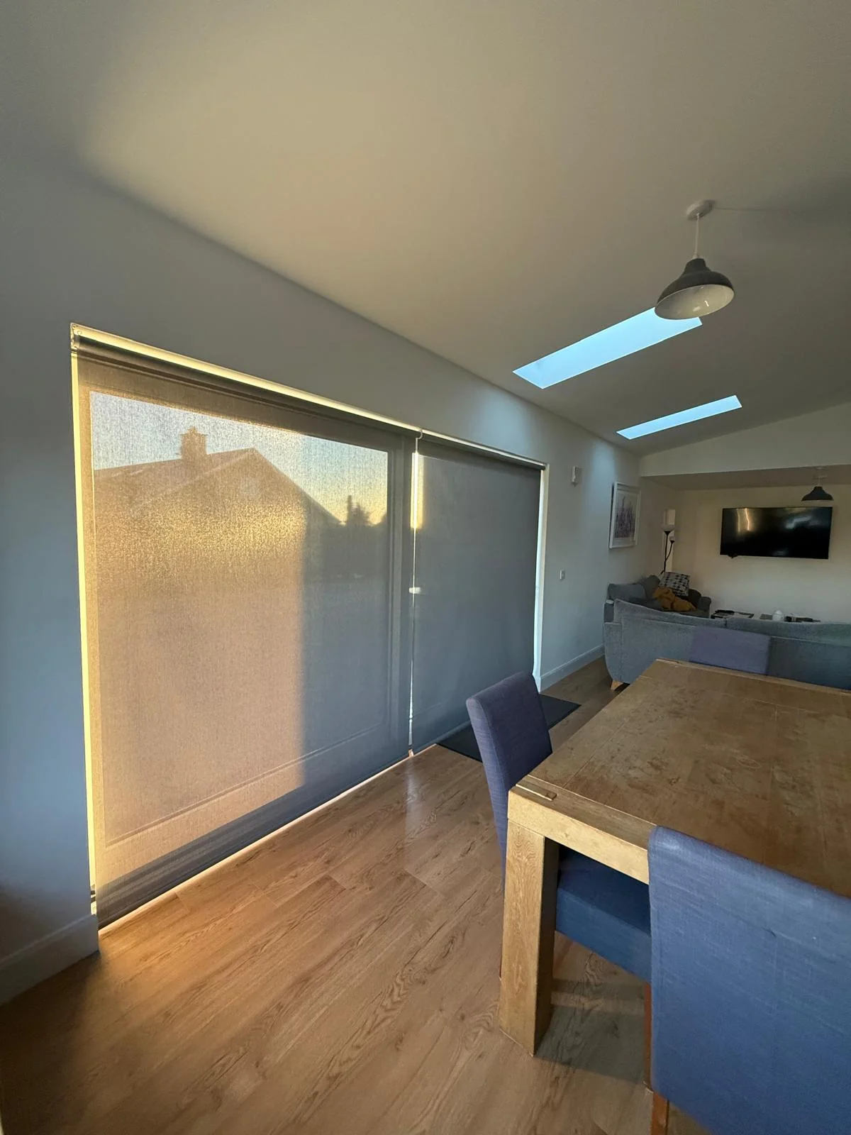 Grey motorised roller blinds filtering low evening sun in a dining and living space